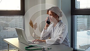 Busy young female doctor in white coat typing on laptop computer and talking on mobile phone.