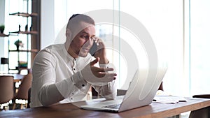 Busy young man is talking on mobile phone and typing on laptop in office at desk