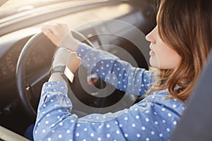 Busy young woman drives car and looks at watch, stuck in traffic jam, hurries to work, being nervous and stressed, feels impatient