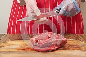 Butcher in red and white apron checking sharpness of his knives in focus. Two freshly cut rib eye steaks on a wooden cutting board