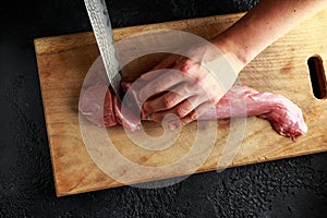 Butcher`s hands ready to cut with a knife raw pork tenderloin fillet on wooden board.