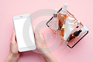 Buy cosmetics online concept. Female hands holding mobile phone with blank screen mockup and shopping basket full of cosmetic