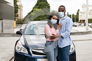 Happy black couple in masks standing near car and hugging