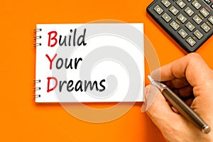 BYD build your dreams symbol. Concept words BYD build your dreams on white note on a beautiful orange background. Businessman hand