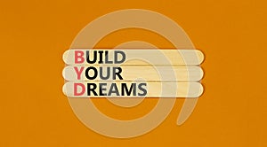 BYD build your dreams symbol. Concept words BYD build your dreams on wooden stick on a beautiful orange table orange background.