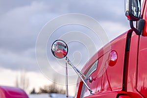 The cab of the red big rig semi truck is reflected in the round rear-view mirror attached to the fender of the truck