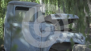 Cabin of broken old truck with green tree leaves around. Side view of abandoned industrial machinery on ruins of