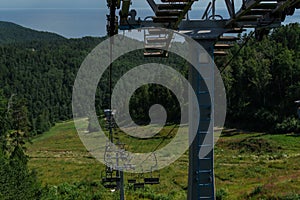 cable car construction with empty seats among green trees, grass field in forest on hill, sunny summer blue sky with white clounds