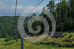 Cable car construction with seats among trees, grass field in green forest on hill, blue sky with white clounds, sea on horizon