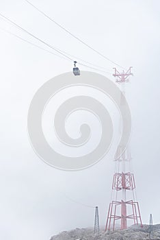 A cable car, mountain gondola suspended on a rope disappearing in the fog. The red-white pylon steel construction is barely visibl