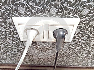 Cables connected to power sockets against a decorative wall. Dual Electrical Outlets With Plugged-In Cables on Patterned