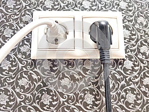 Cables connected to power sockets against a decorative wall. Dual Electrical Outlets With Plugged-In Cables on Patterned