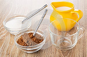 Cacao powder, spoon in bowl, teaspoon in bowl with sugar, pitcher with milk, cup on wooden table