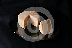 Caciotta cheese on a black plate. A piece of triangular shape is cut off from the cheese. Black background. Isolated. Close-up