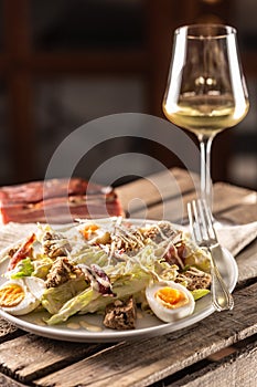 Caesar salad with boiled eggs, bacon, and croutons served on a plate next to a glass of white wine