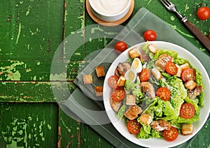 Caesar salad with chicken fillet, tomatoes, croutons and parmesan in a plate on a rustic wooden background, top view