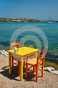 Cafe tableon beach in Adamantas town on Milos island with Aegean sea with boats in background