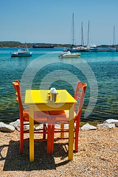 Cafe tableon beach in Adamantas town on Milos island with Aegean sea with boats in background