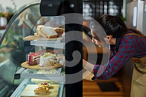 Cafe worker is opening bakery cabinet, bent down use tongs pick cake in refrigerator out put them on sall tray, that is served