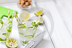 Caipirinha, mojito cocktail with lime, brown sugar, ice and mint leaves in beautiful glasses, cut green citrus on white wooden bac