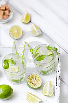 Caipirinha, mojito cocktail with lime, brown sugar, ice and mint leaves in beautiful glasses, cut green citrus on white wooden bac