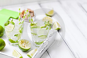 Caipirinha, mojito cocktail with lime, brown sugar, ice and mint leaves in beautiful glasses, cut green citrus on white wooden bac