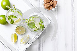 Caipirinha, mojito cocktail with lime, brown sugar, ice and mint leaves in beautiful glasses, cut green citrus on white wooden bac