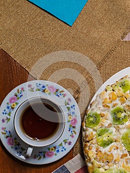 A cake made from biscuits, decorated with kiwi slices, lies in a white plate next to a saucer and a cup of black tea on checkered