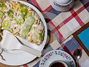 A cake made from biscuits, decorated with kiwi slices, lies in a white plate next to a saucer and a cup of black tea on checkered