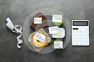 Calculator and food products with calorific value tags on dark grey table, flat lay. Weight loss concept photo