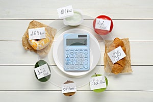 Calculator and food products with calorific value tags on white wooden table, flat lay. Weight loss concept