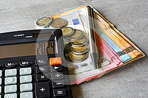 Calculator with number 2024 on display, pen, euro banknotes and coins on grey background