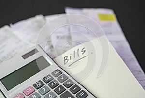 Calculator on top of notepad with the words Bills. Stack of bills at the background