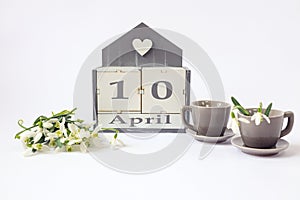 Calendar for April 10: cubes with the number 10, the name of the month in English, two gray coffee cups, a bouquet of snowdrops on