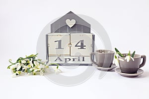 Calendar for April 14: cubes with the number 14, the name of the month in English, two gray coffee cups, a bouquet of snowdrops on