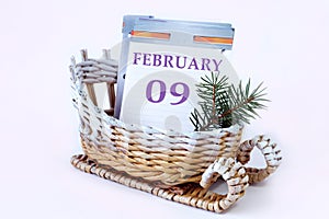 Calendar for February 9: decorative sleigh with a loose-leaf calendar inside, name of the month in English, numbers 09, fir branch