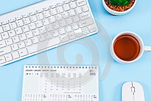 Calendar for January 2022 with keyboard, mouse, mug of tea and small flower pot on light blue