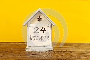 Calendar for November 24: the name November in English, numbers 24 on a decorative house on a wooden table, yellow background