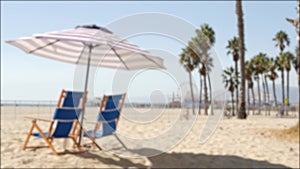 California beach defocused, two empty blue deck chairs, striped umbrella near pier in Santa Monica pacific ocean resort