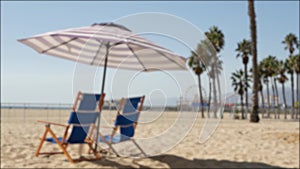 California beach defocused, two empty blue deck chairs, striped umbrella near pier in Santa Monica pacific ocean resort