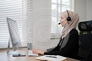 Call center worker, young Muslim woman wearing hijab, talking to customer on call phone on computer in customer service