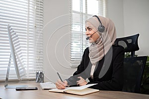 Call center worker, young Muslim woman wearing hijab, talking to customer on call phone on computer in customer service