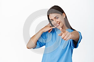 Call me. Smiling modern girl pointing finger at camera and showing phone gesture, inviting you, standing over white
