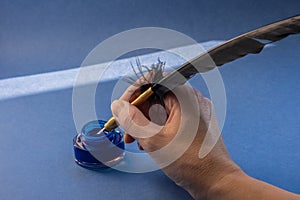 Calligraphic blue ink in a glass inkwell, hand holding a classic black quill pen with a golden metallic nib, deep blue background