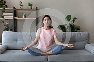 Calm Arabian woman meditating, sitting in lotus pose on couch