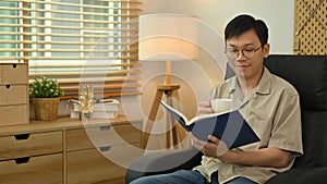 Calm asian man wearing glasses sitting on sofa and reading book. Leisure and people concept