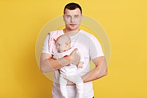 Calm attractive single father dresses white t shirt posing together with newborn daughter isolated over yellow background, man