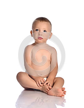 Calm baby boy toddler in diaper sits on the floor, touching his feet and looks at upper corner