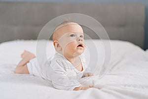 Calm beautiful smiling infant baby lying on bed and looking away. Curious sweet newborn kid in white costume lying on