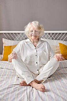 calm caucasian senior woman engaged in yoga, sit on bed with crossed legs